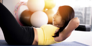 Close-up of young active and fitness Asian woman doing sit ups and crunches inside gym with exercise ball in background