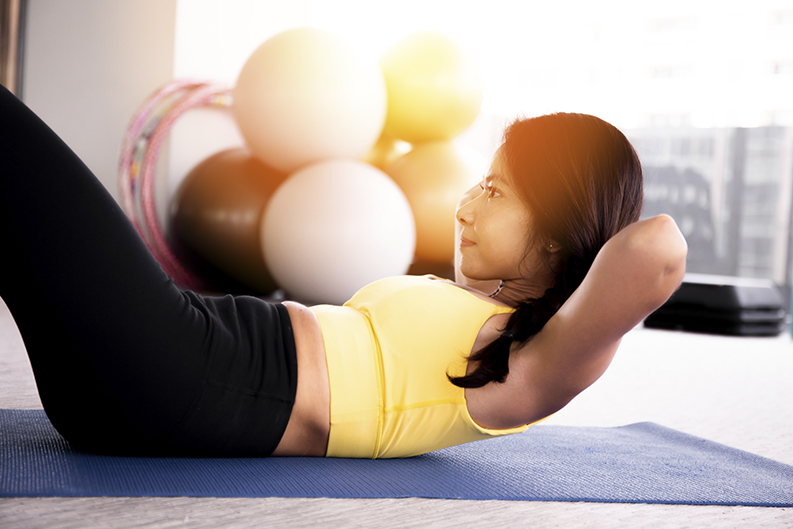 Close-up of young active and fitness Asian woman doing sit ups and crunches inside gym with exercise ball in background